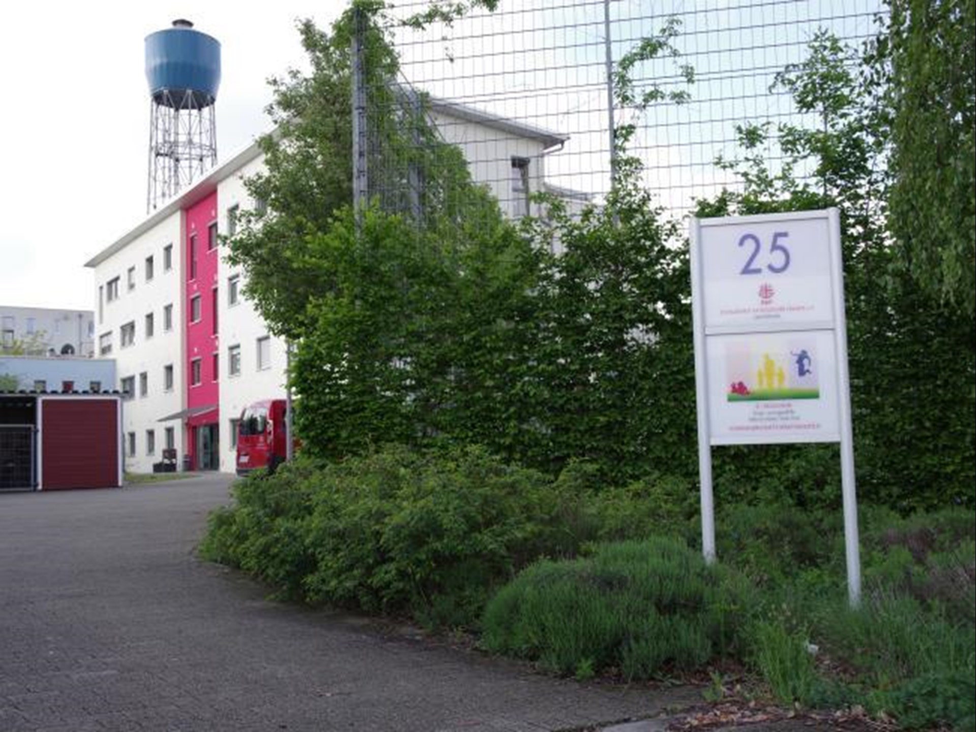 Ein Wohngebäudeanlage mit einem Schild, das die Nummer 25 anzeigt.  Die Szene zeigt ein Gebäude mit einem roten Streifen und einem blauen Wasserturm.  Es gibt viel Grün um
